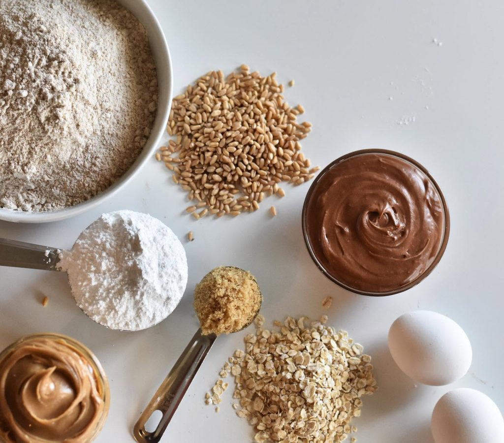 Ingredients laid out: flour, wheat berries, oats, peanut butter, chocolate and eggs